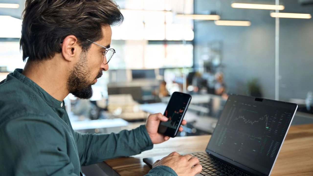 Trader holding a phone in front of a laptop, analyzing forex charts to illustrate the concept of a moving average crossover in trading.